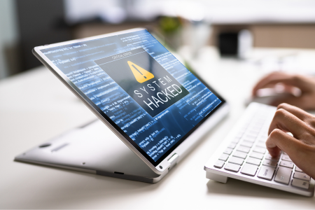 A laptop screen displaying a "System Hacked" warning with lines of code in the background, symbolizing a cyberattack. Hands are typing on a keyboard in the foreground.